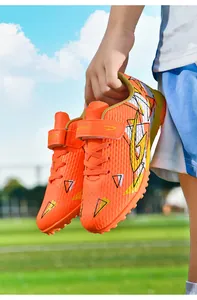 Oranje voetbalschoenen voor kinderen met klittenband op een zonnig kunstgrasveld.
