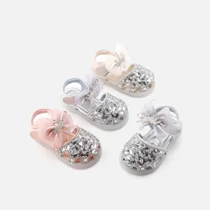 Four pairs of baby girl princess sandals with sequins, crowns, and bows on grey background