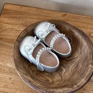 Zapatos Mary Jane plateados para niña con perlas y lazo, presentados en un cuenco de madera.