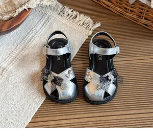 Close-up of silver kids sandals with lace bow and floral charms on a textured woven mat