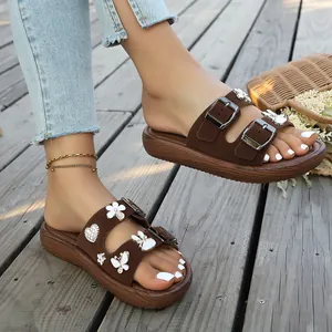 Nahaufnahme brauner Plateausandalen mit Strassherz- und Schmetterlingsanhängern auf Holzdeck.