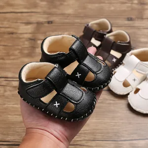 Hand hält ein Paar schwarze Babysandalen aus Kunstleder mit weißer Kreuzstickerei