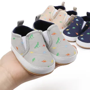 Hand holding cute dinosaur print baby slip-on shoes for newborns 0-1 year old.