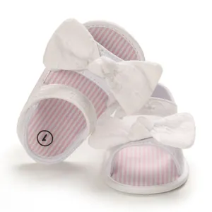 Primo piano di sandali bianchi per bambina con strisce rosa e grande fiocco floreale.