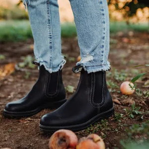 Primer plano de botas Chelsea de cuero negro en un entorno de jardín con frutas caídas.