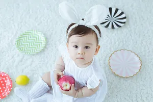 Adorable baby girl in white lace dress and bunny headband playing with a red apple