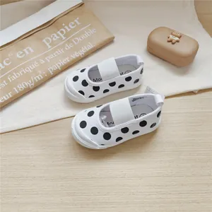 White canvas baby shoes with black polka dots and elastic strap on wooden surface.