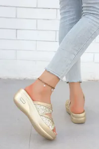 Dettaglio di sandali con plateau glitterati oro e cavigliera di perle contro un muro bianco.