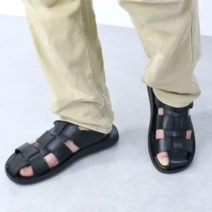 Close-up of men's black leather Roman sandals worn with khaki trousers on gray floor.