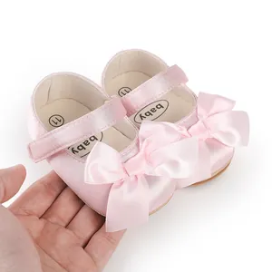 Detail view of pink satin baby walking shoes with a decorative bow held by hand