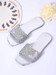 Dettaglio di sandali piatti glitter argento con un accento decorativo a farfalla.