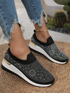 Primo piano di sneakers nere con zeppa e strass indossate su un tappeto di paglia.