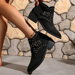 Detalle de botines negros de punta con tachuelas y correa con hebilla sobre tacón de bloque.