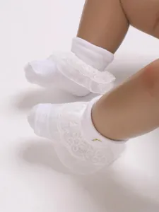 Close-up detail of white baby socks featuring delicate lace ruffles and a gold cross