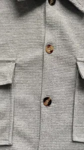 Close-up detail of grey textured shirt fabric featuring amber buttons and flap pockets.