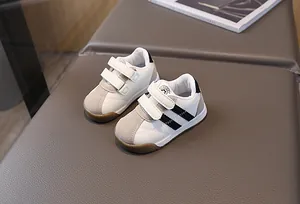 Detailed view of white toddler sneakers with black stripes and velcro straps on a soft bench.