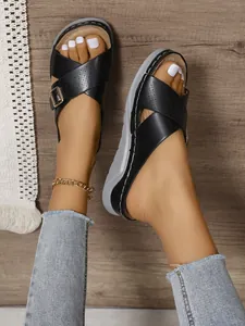 Close-up of black wedge sandals with cross straps and metal buckle on wood floor.