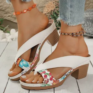 Detail of white floral block heel sandals worn with elegant anklets on wooden deck.