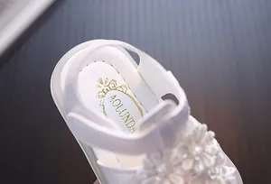 Nahaufnahme weißer Kleinkindsandale mit Spitzenblumendetail und goldenem Markenlogo.