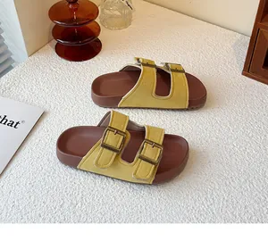 Gros plan de claquettes en daim jaune avec doubles boucles en laiton sur surface blanche.