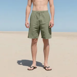 Male model wearing olive green linen drawstring casual shorts on a sunny minimalist beach