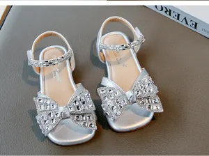 Détail de sandales argentées pour filles avec nœuds en strass brillants et bout carré.