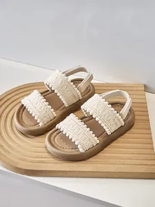 Primer plano de sandalias para niña estilo étnico con encaje de ganchillo y pompones beige.