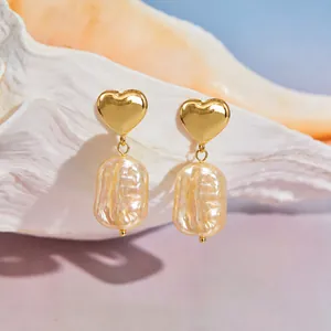 Vue détaillée de boucles d'oreilles cœur dorées avec perles baroques posées sur un coquillage.