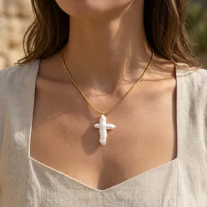 Gros plan d'un pendentif croix en perle baroque sur une élégante chaîne serpent dorée.