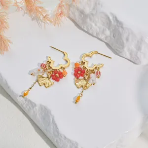 Close-up of gold ginkgo leaf earrings with red bead flowers and tassels on a white stone background.