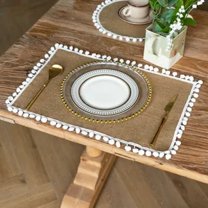 Close-up detail of a rustic jute placemat with white pom-pom fringe on a wooden table setting