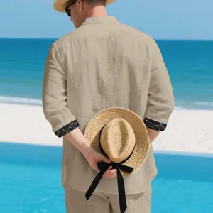Back view of a man in a beige linen shirt set on a sunny beach holding a straw hat