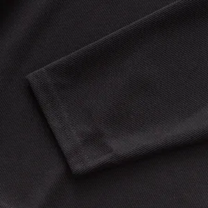 Détail macro du poignet en maille côtelée française sur un polo noir à manches longues.
