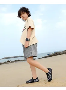 Child wearing black and grey checkered water shoes on a sandy beach with ocean background