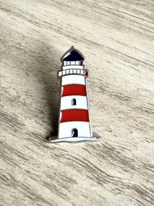 Détail d'un pin's en émail en forme de phare avec des rayures rouges et blanches vives.