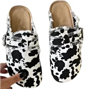 Sabot da donna con stampa mucca, dettaglio frange e cinturino con fibbia su fondo bianco.