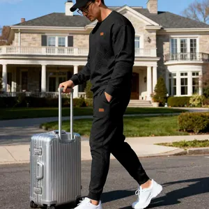 Man in black jacquard tracksuit pulling a silver suitcase in front of a house