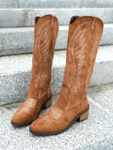 Macro view showing detailed white embroidery on brown suede Western cowboy high boots.