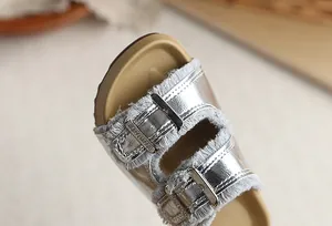 Primer plano de sandalias plateadas para niña con hebillas y bordes deshilachados.