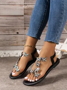 Primer plano de sandalias planas negras con mariposas de pedrería en tiras transparentes.