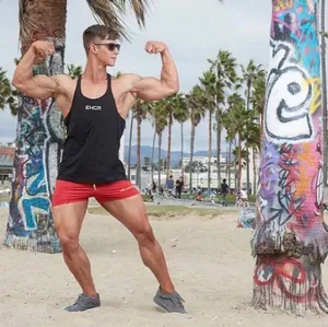 Muscular man wearing a black fitness tank top posing at a sunny beach with palm trees