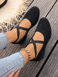 Dettaglio di scarpe zeppa in camoscio nero con cinturini elastici incrociati su legno.