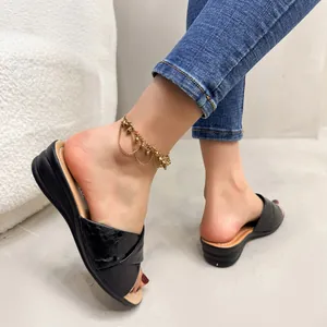 Black leather wedge slippers with crossover straps shown worn on feet with a gold anklet.