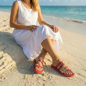 Red and gold flat casual sandals worn by a woman sitting on a sunny sandy beach scene