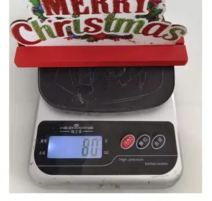 Detail shot of a red and white wooden Merry Christmas sign weighing 80 grams on a digital scale.