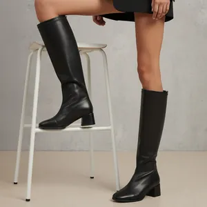 Side view of woman wearing black square toe block heel knee-high boots on stool