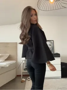 Mujer elegante con chaqueta de capa negra y cinturón con hebilla metálica en un dormitorio