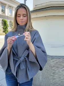 Detalle de una chaqueta tipo capa gris con botón metálico dorado y cinturón de lazo