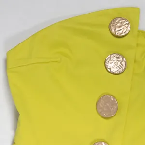 Detailansicht der großen goldgeprägten Knöpfe an einem lindgelben trägerlosen Jumpsuit.
