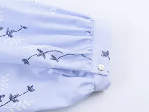 Detail van een blauwe geborduurde hemdmanchet met bloemmotieven en een witte knoop.